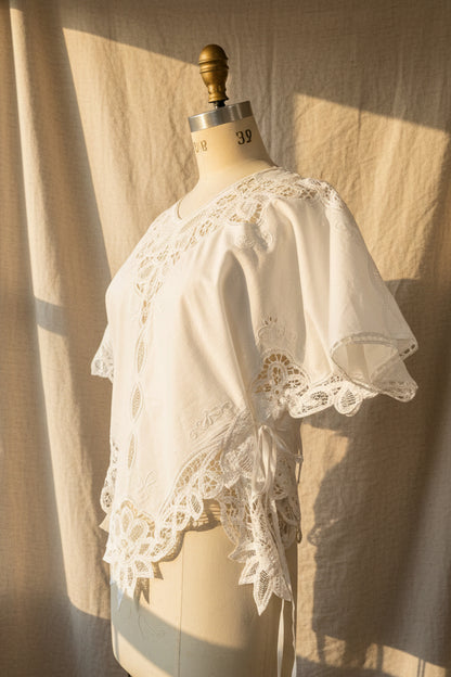 A white embroidered lace-trim blouse displayed on a cream dress form mannequin, with a neutral indoor room background.