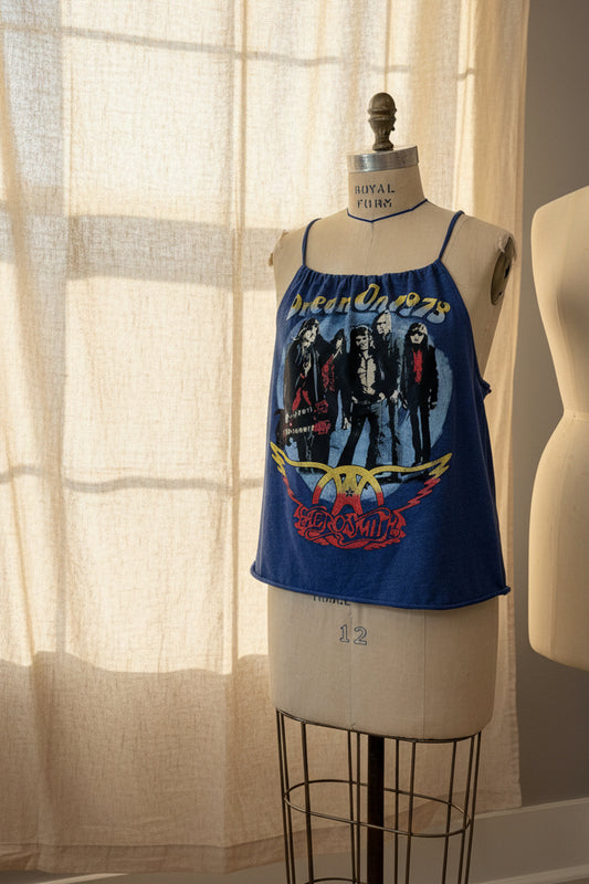 A blue sleeveless graphic tank top with spaghetti straps, featuring a heavy metal band print, displayed on a cream-colored dress form mannequin in front of a beige curtain.