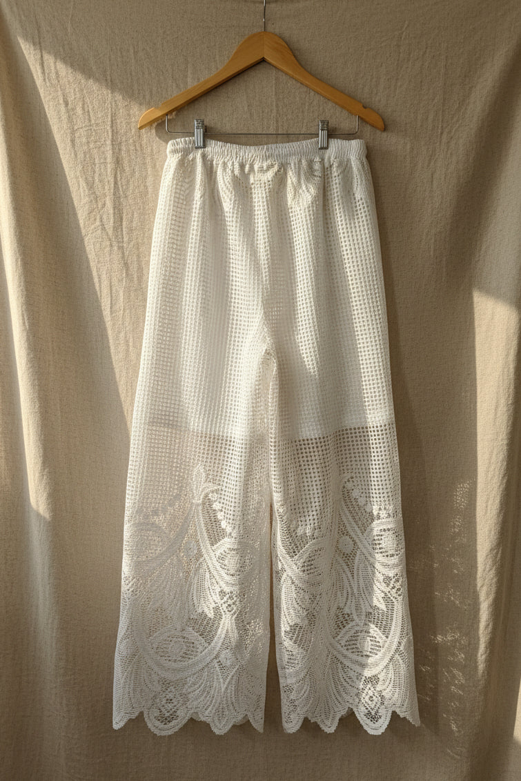 White crochet knit cropped pants with a scalloped lace hem, hanging on a wooden hanger against a white paneled door.