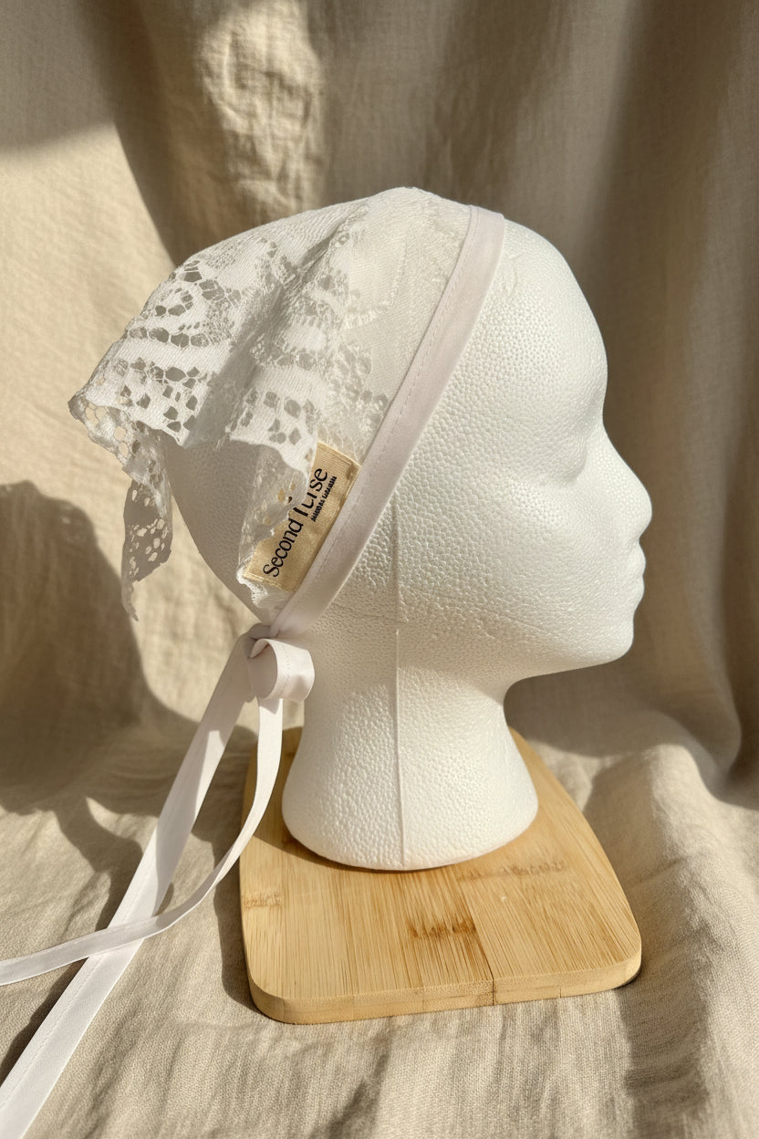 An off-white lace bonnet with a matching ribbon tie, displayed on a white styrofoam mannequin head resting on a small wooden stand. A small paper tag on the bonnet reads 'Second Verse'.