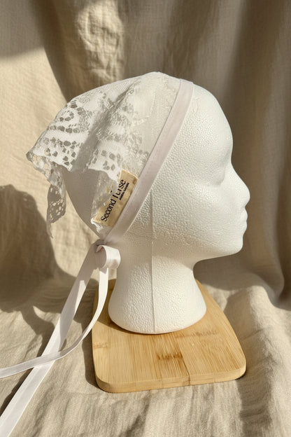 An off-white lace bonnet with a matching ribbon tie, displayed on a white styrofoam mannequin head resting on a small wooden stand. A small paper tag on the bonnet reads 'Second Verse'.