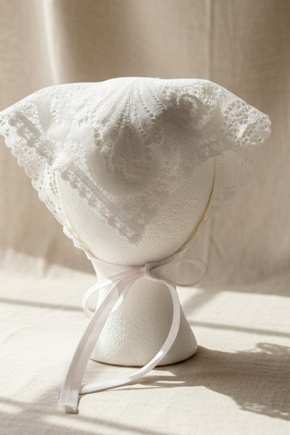White lace bonnet with a white ribbon tie, displayed on a white mannequin head stand resting on a small wooden board.
