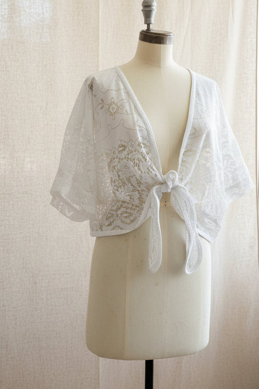 White lace short-sleeve wrap top with a tie-front detail, displayed on a cream dress form mannequin in an indoor room setting.