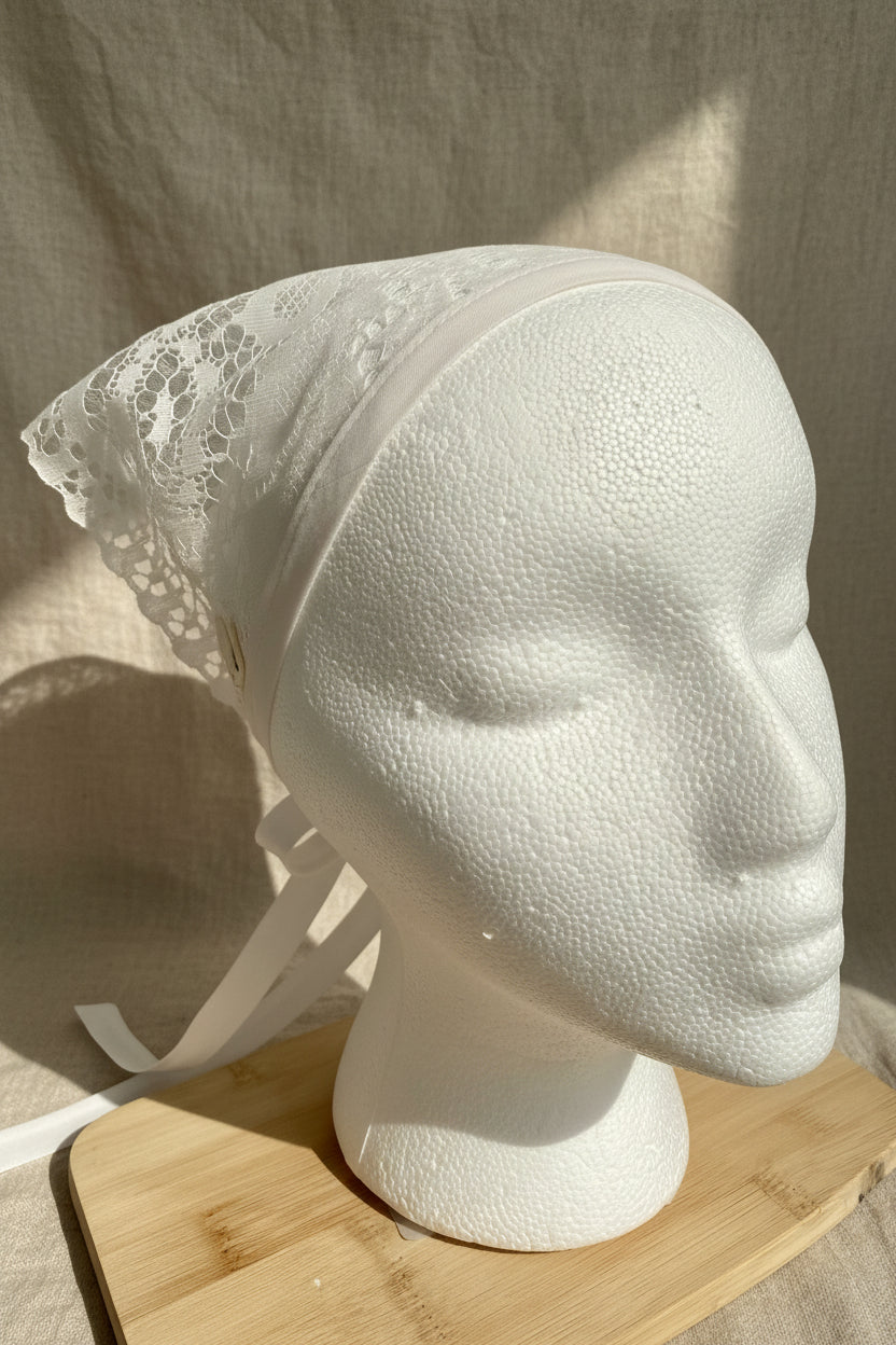 White lace head covering with a matching ribbon, displayed on a white styrofoam mannequin head resting on a wooden board.