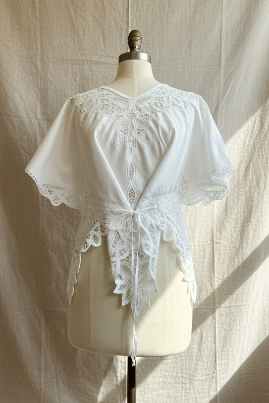 White short-sleeve lace top displayed on a cream dress form mannequin, featuring intricate floral lace applique around the neckline, sleeves, and hem, with a lace tie at the waist.
