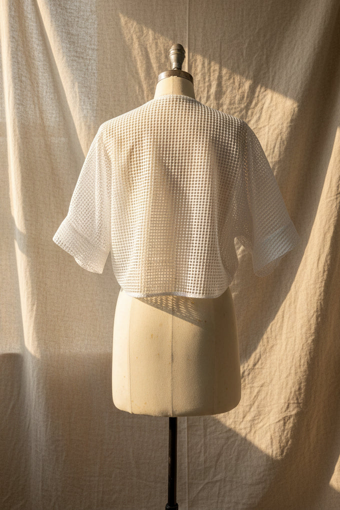 White open-knit short-sleeve cropped top displayed on a cream dress form, photographed indoors against a light gray wall and white door.