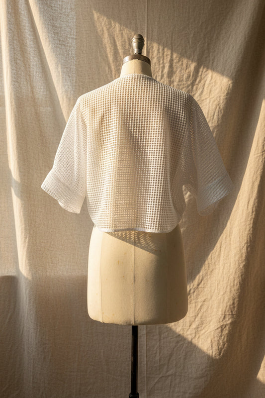 White open-knit short-sleeve cropped top displayed on a cream dress form, photographed indoors against a light gray wall and white door.