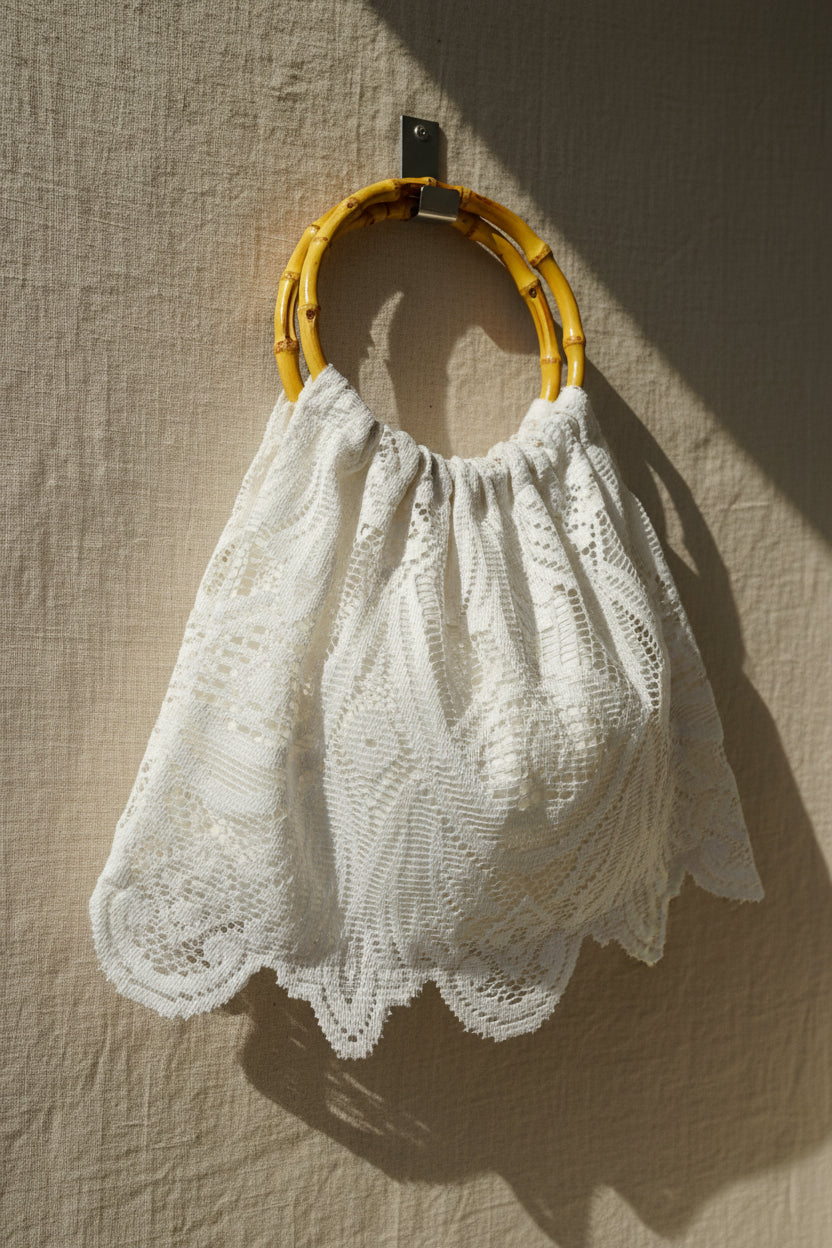 A white crocheted lace handbag with a scalloped edge and a natural bamboo ring handle, hanging against a white paneled door.