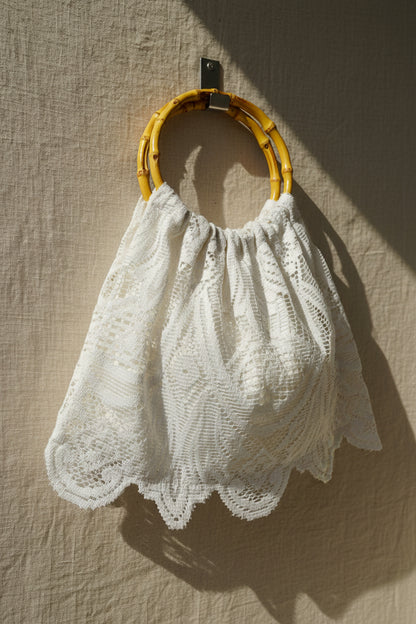 A white crocheted lace handbag with a scalloped edge and a natural bamboo ring handle, hanging against a white paneled door.