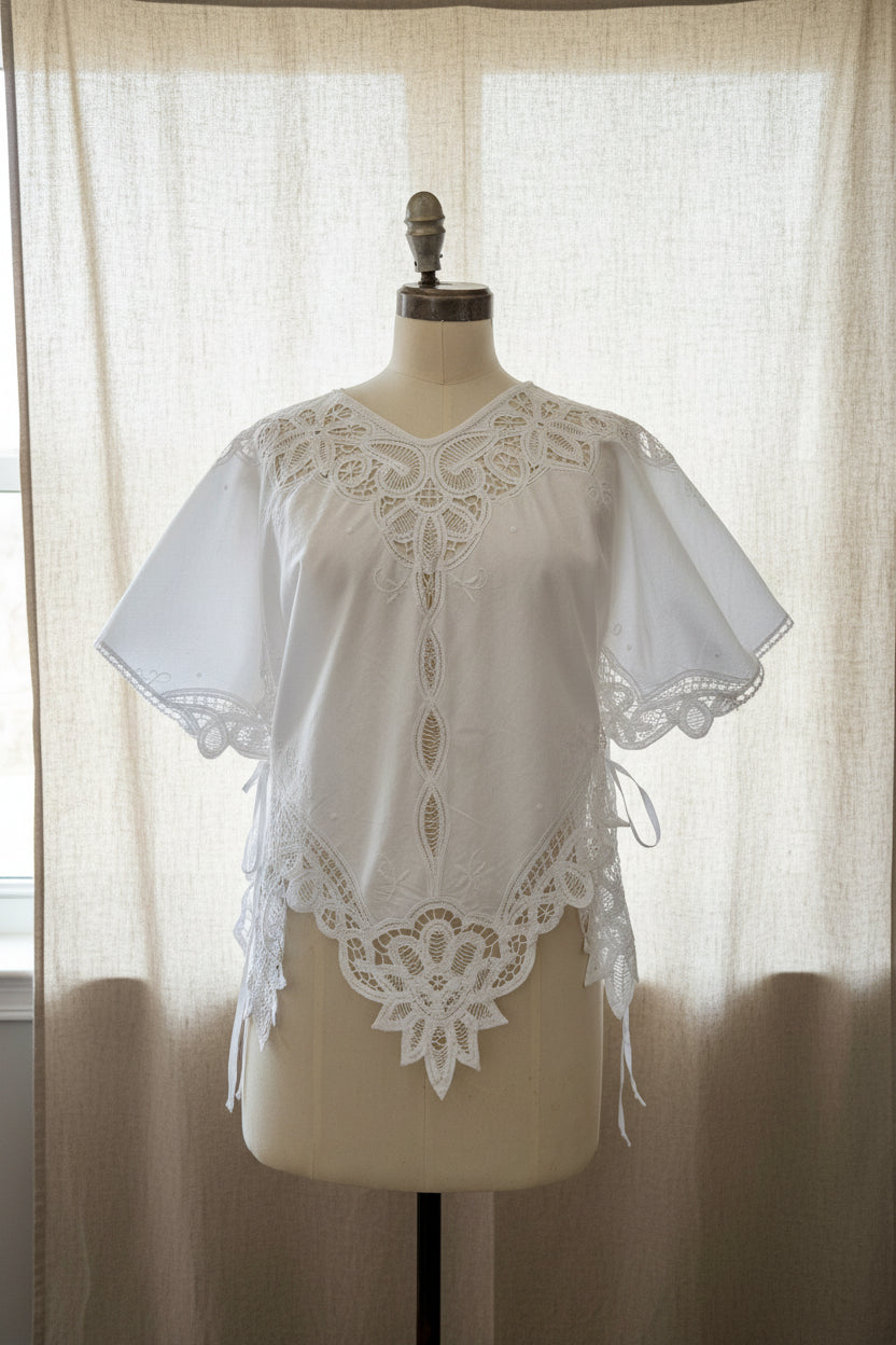 White embroidered blouse with lace trim and short flutter sleeves, displayed on a cream dress form in an indoor room.