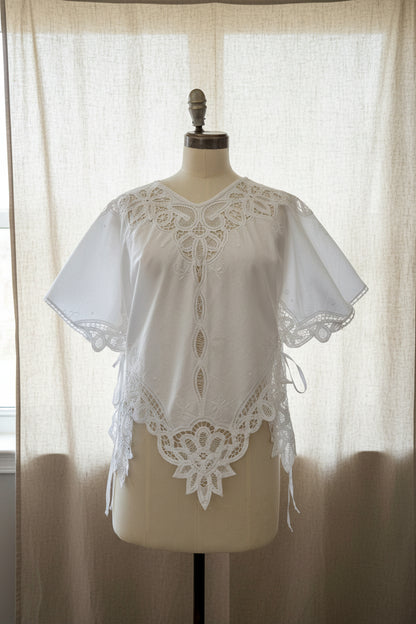 White embroidered blouse with lace trim and short flutter sleeves, displayed on a cream dress form in an indoor room.