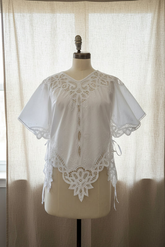 White embroidered blouse with lace trim and short flutter sleeves, displayed on a cream dress form in an indoor room.