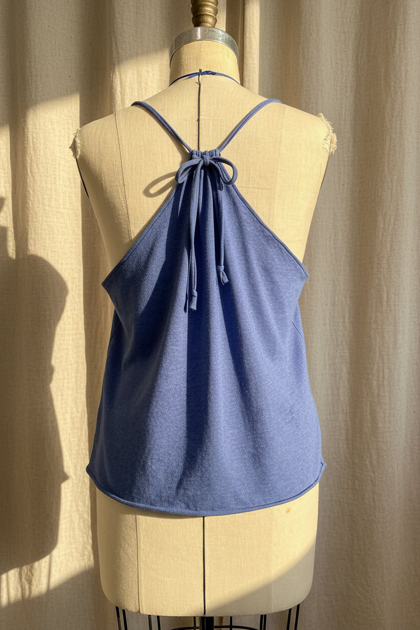 A dusty blue halter-style camisole top displayed on a cream-colored dress form mannequin, set against a draped beige fabric backdrop with warm sunlight and shadows.