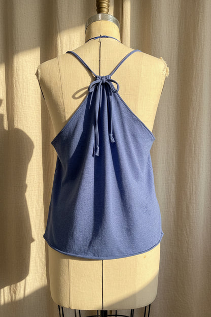 A dusty blue halter-style camisole top displayed on a cream-colored dress form mannequin, set against a draped beige fabric backdrop with warm sunlight and shadows.