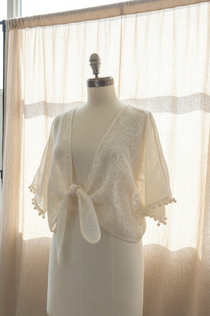 Mannequin wearing a white lace shrug in front of sheer curtains