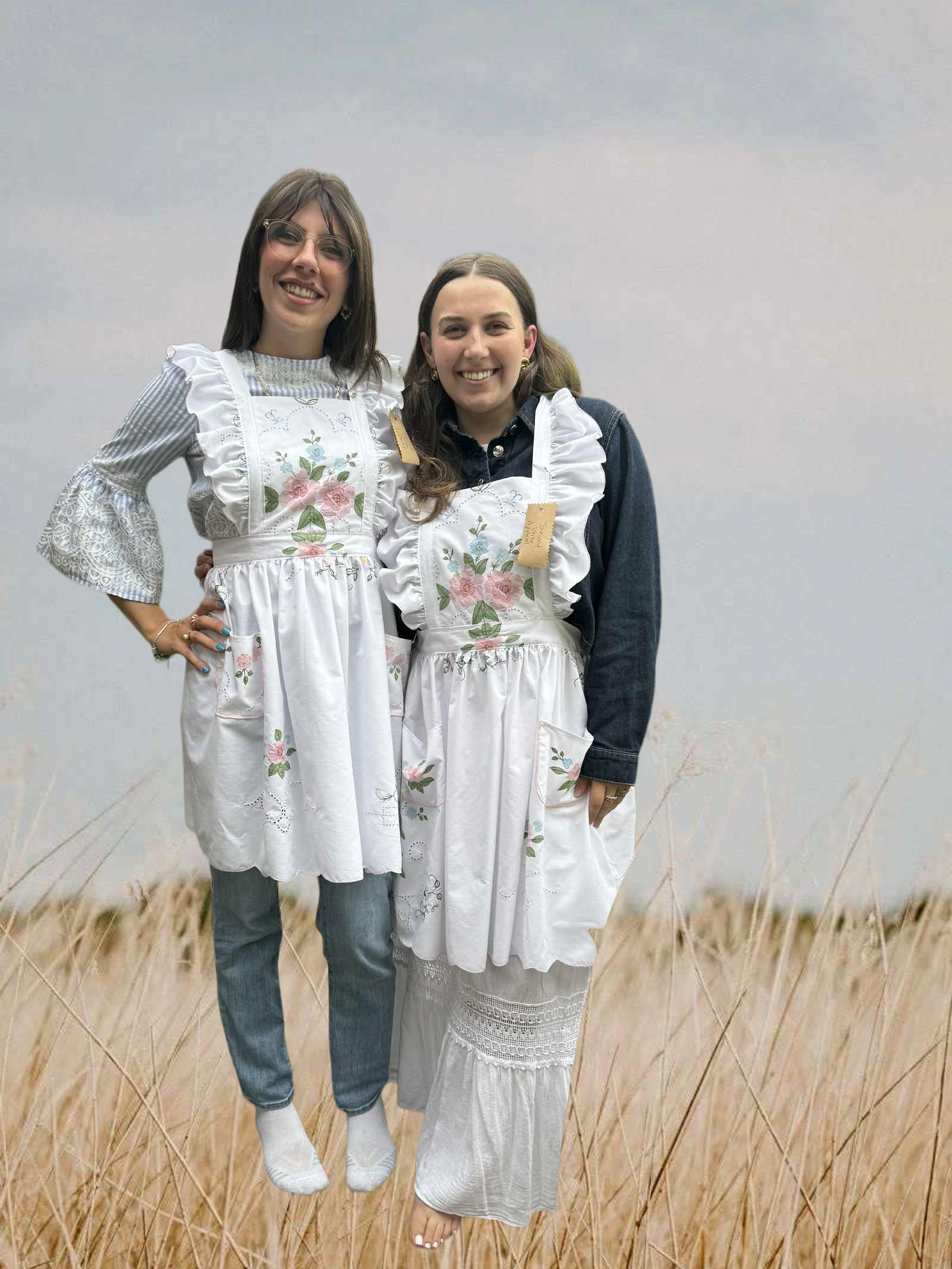 Two women in matching white 1950's style aprons  with floral embroidery standing in a field.