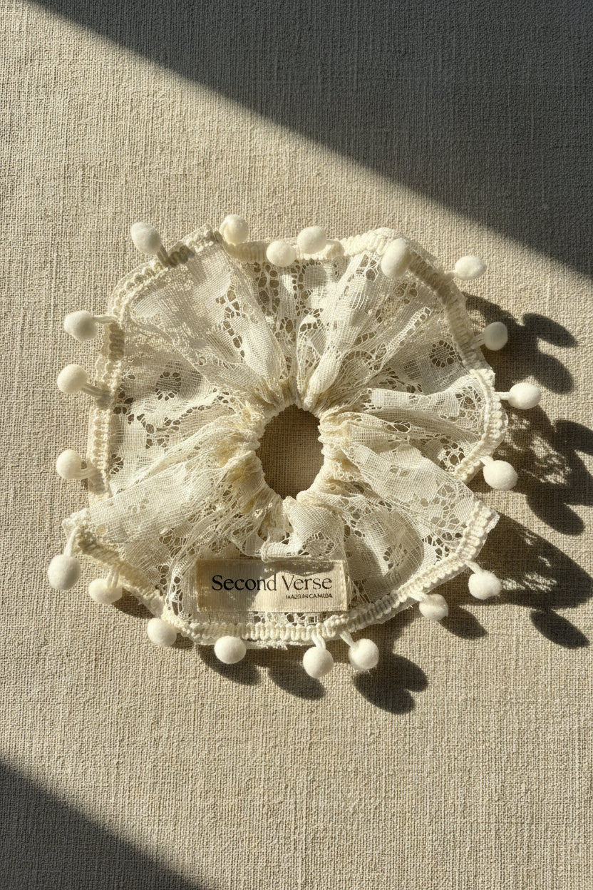 Ivory lace hair scrunchie with pom-poms on a beige surface