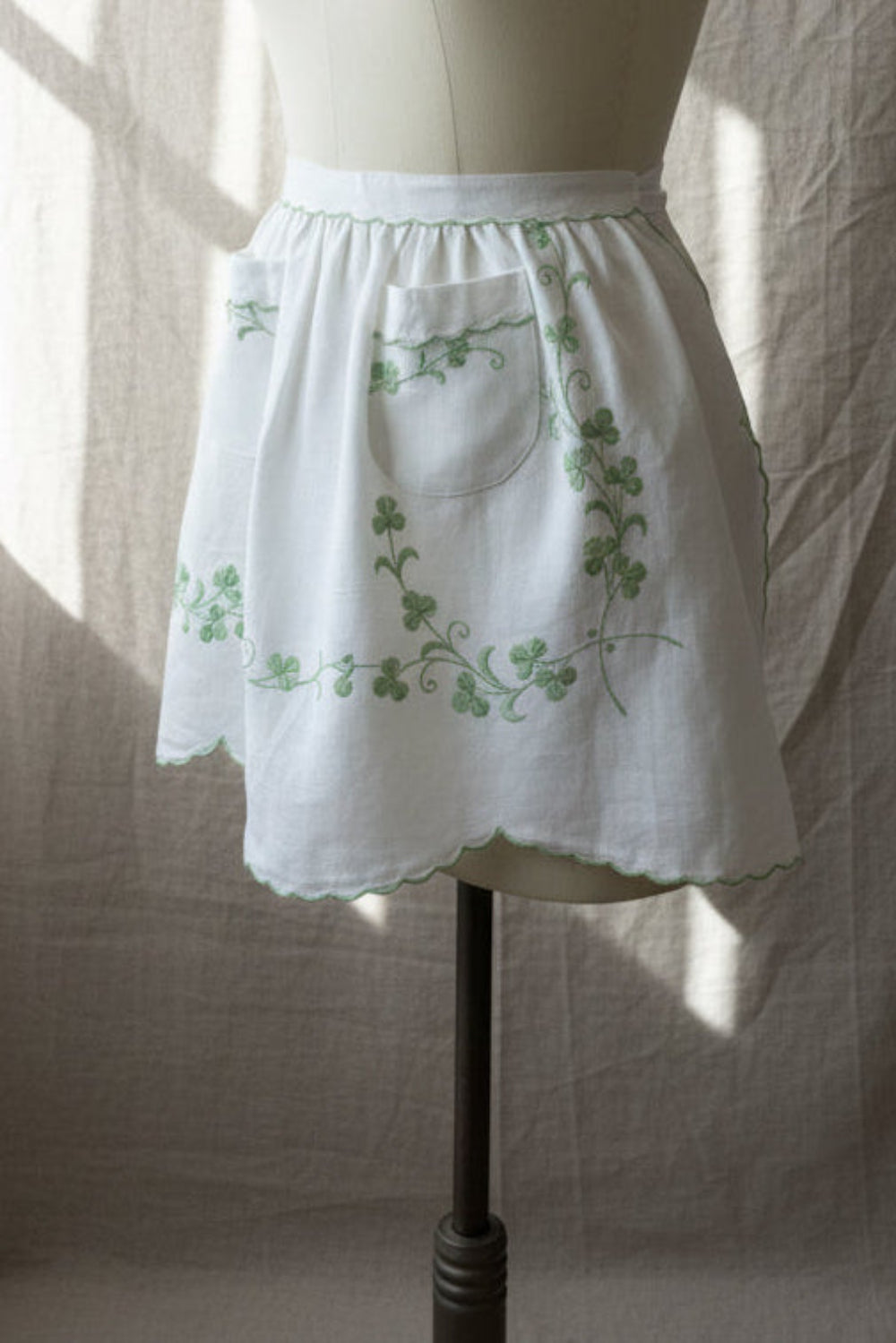 White apron with green embroidery on a mannequin against a neutral background