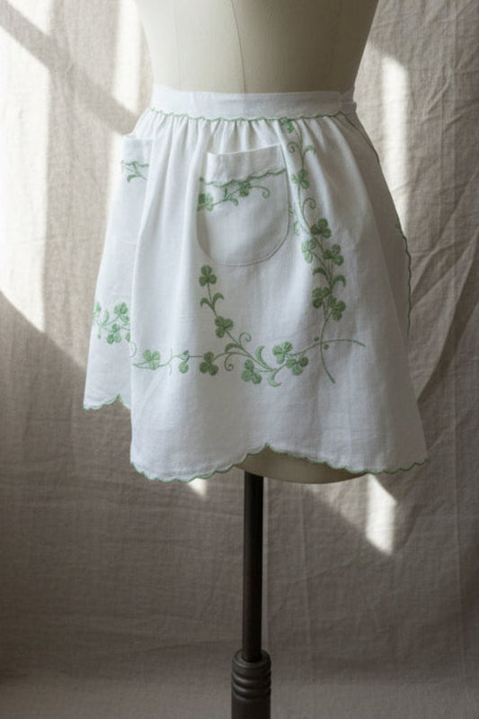 White apron with green embroidery on a mannequin against a neutral background