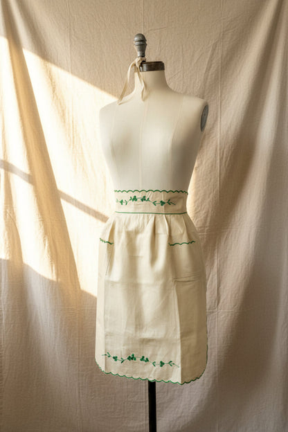 Ivory apron with green embroidery on a mannequin against a white wall.